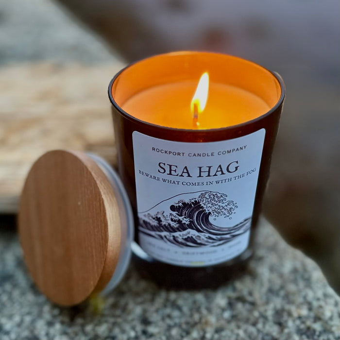 A lit black glass candle jar labeled 'Sea Hag' by Rockport Candle Company, with a wooden lid resting beside it on a granite surface.